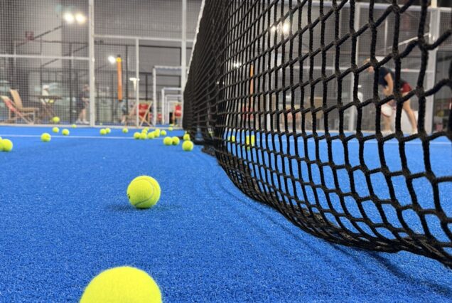 Manche de tennis en gazon synthétique bleu avec plusieurs balles jaunes, vue rapprochée, salle d'entraînement de padel avec filet et pratiquants en arrière-plan, ambiance sportive et dynamique.