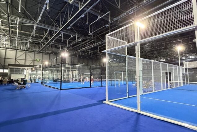 Court de padel intérieur avec sol en tapis bleu, structure en verre et cage de protection en métal, ambiance moderne pour pratique sportive.