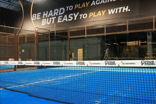 TEXTE-ALT : Court de padel intérieur avec surface en bleu, entouré d'une clôture métallique, éclairage puissant, grand panneau motivant, logo "Your Padel" visible au sol.